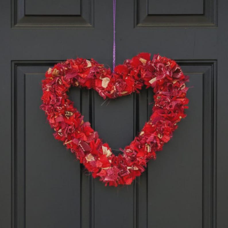 May include: A heart-shaped wreath made of red fabric scraps hangs against a dark door. The wreath is a vibrant mix of red hues, with a textured appearance. A purple string is used for hanging, adding a subtle contrast to the overall design.