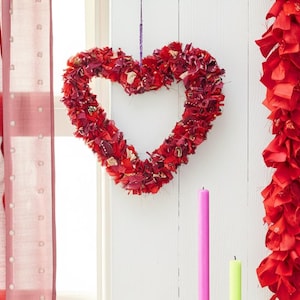 May include: A red heart-shaped wreath made of fabric hangs on a white wall. A red garland and two candles, one pink and one green, are also visible. The wreath is suspended by a purple string. The scene is lit by natural light.