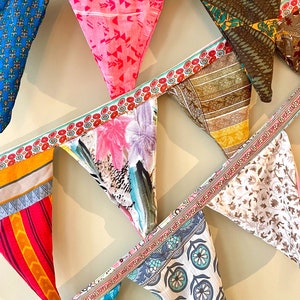 May include: A colorful fabric bunting with triangular flags featuring various patterns, including floral, geometric, and abstract designs. The flags are connected by a white ribbon with a purple and blue trim.
