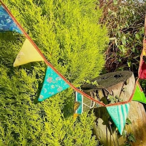 May include: A colorful fabric bunting with triangular flags in shades of blue, yellow, green, and red. The bunting is strung between a tree and a wooden stump.