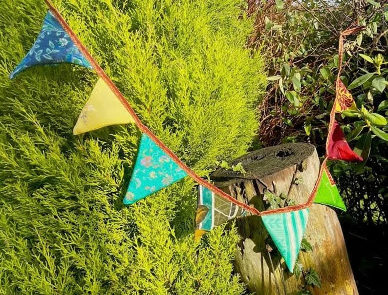 May include: A colorful fabric bunting with triangular flags in shades of blue, yellow, green, and red. The bunting is strung between a tree and a wooden stump.