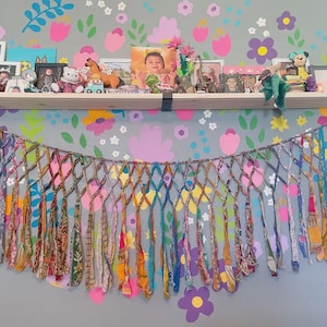 Puede incluir: Un colorido tapiz de pared textil hecho a mano con un diseño de red en forma de diamante. El tapiz presenta largas tiras verticales de tela en varios colores y patrones, creando una estética vibrante y bohemia. Una estantería encima muestra fotos enmarcadas y objetos decorativos.