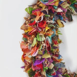 May include: A colorful, handmade wreath composed of layered fabric scraps in various colors, including red, green, purple, and yellow. The wreath is set against a white background, showcasing its vibrant, textured design. This decorative piece is suitable for seasonal home decor.