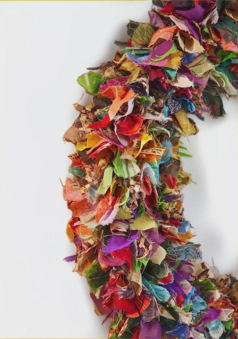 May include: A colorful, handmade wreath composed of layered fabric scraps in various colors, including red, green, purple, and yellow. The wreath is set against a white background, showcasing its vibrant, textured design. This decorative piece is suitable for seasonal home decor.