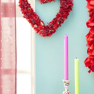 May include: A red fabric heart-shaped wreath hangs on a light blue wall, with two candles in decorative holders. A pink and a yellow candle stand upright. A red curtain is on the left.