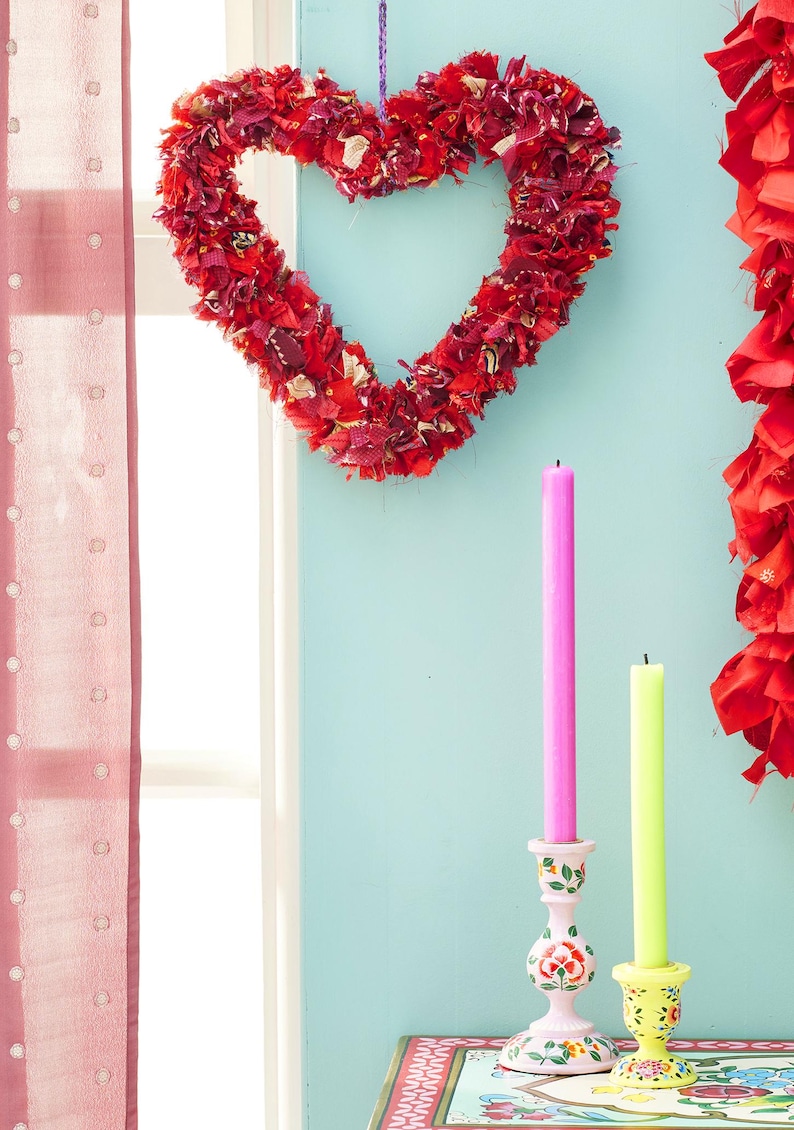 May include: A red fabric heart-shaped wreath hangs on a light blue wall, with two candles in decorative holders. A pink and a yellow candle stand upright. A red curtain is on the left.