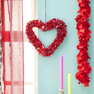 May include: A heart-shaped wreath made of red fabric scraps hangs against a light blue wall. A red fabric garland and two candles, one pink and one yellow, are also visible. The scene is set near a window with red curtains.