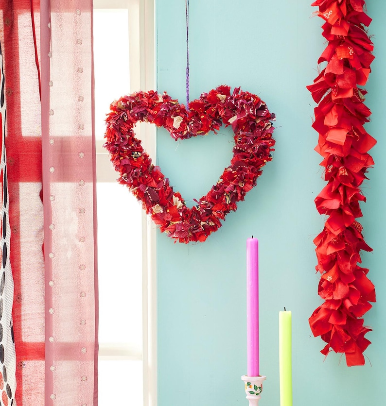 May include: A heart-shaped wreath made of red fabric scraps hangs against a light blue wall. A red fabric garland and two candles, one pink and one yellow, are also visible. The scene is set near a window with red curtains.