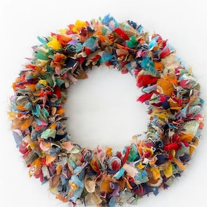 May include: A colorful, handmade wreath composed of fabric scraps in various colors, including red, blue, yellow, and orange. The wreath is circular and has a dense, textured appearance, perfect for home decor.