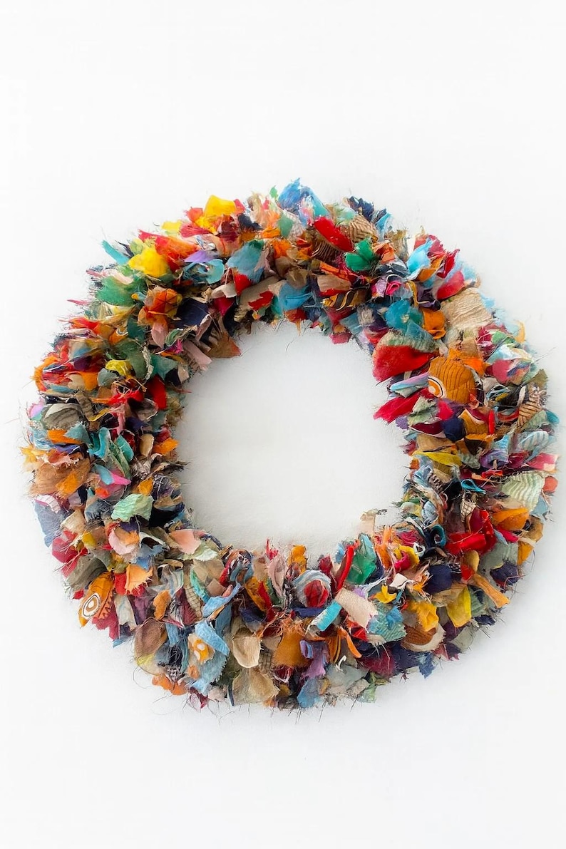 May include: A colorful, handmade wreath composed of fabric scraps in various colors, including red, blue, yellow, and orange. The wreath is circular and has a dense, textured appearance, perfect for home decor.