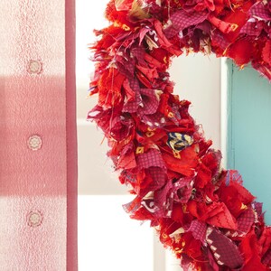 May include: A heart-shaped wreath made of layered red fabric scraps. The wreath features various shades of red and burgundy fabrics, creating a textured and vibrant appearance. The wreath is displayed against a light blue wall and a pink curtain.