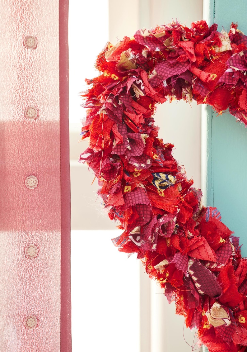 May include: A heart-shaped wreath made of layered red fabric scraps. The wreath features various shades of red and burgundy fabrics, creating a textured and vibrant appearance. The wreath is displayed against a light blue wall and a pink curtain.