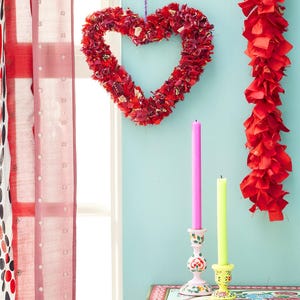 May include: A red heart-shaped wreath made of fabric hangs against a light blue wall. A red garland and two candlesticks with pink and yellow candles are also visible. A patterned table and red curtains complete the scene.