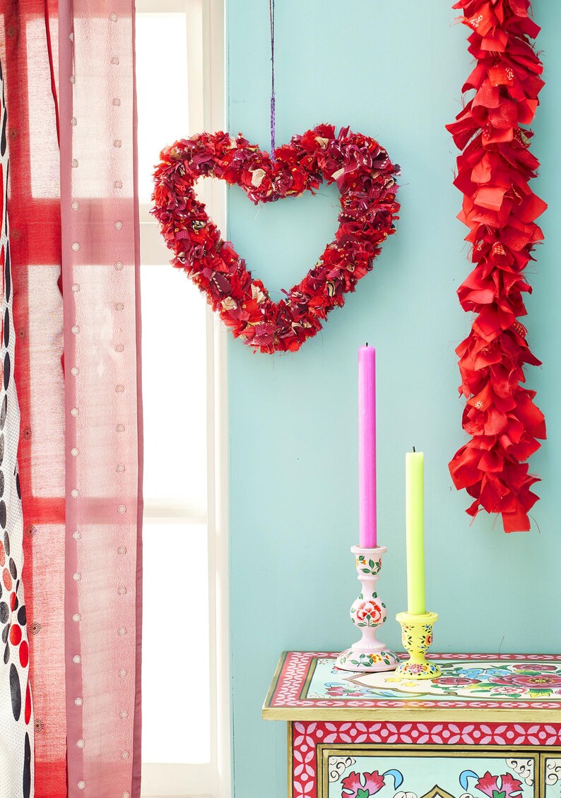 May include: A red heart-shaped wreath made of fabric hangs against a light blue wall. A red garland and two candlesticks with pink and yellow candles are also visible. A patterned table and red curtains complete the scene.