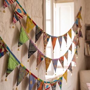 Pode incluir: Uma grinalda decorativa com duas filas de bandeiras triangulares de tecido em várias cores e padrões, incluindo verde, amarelo, azul e vermelho. Cada bandeira individual é rematada com um pequeno sino de metal dourado pendurado.