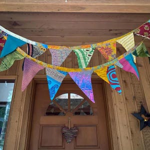 Könnte beinhalten: Ein farbenfrohes Banner aus dreieckigen Flaggen hängt über einer Holztür. Die Flaggen weisen verschiedene Muster und Farben auf, darunter Blau, Rosa, Gelb und Grün. Unter dem Türgriff hängt eine herzförmige Dekoration aus geflochtenem Material.