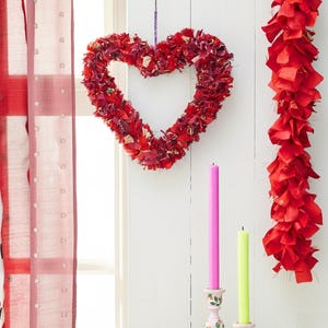 May include: A red heart-shaped wreath made of fabric hangs on a white wall. A red fabric garland is to the right. Two candles, one pink and one green, sit in decorative holders below.
