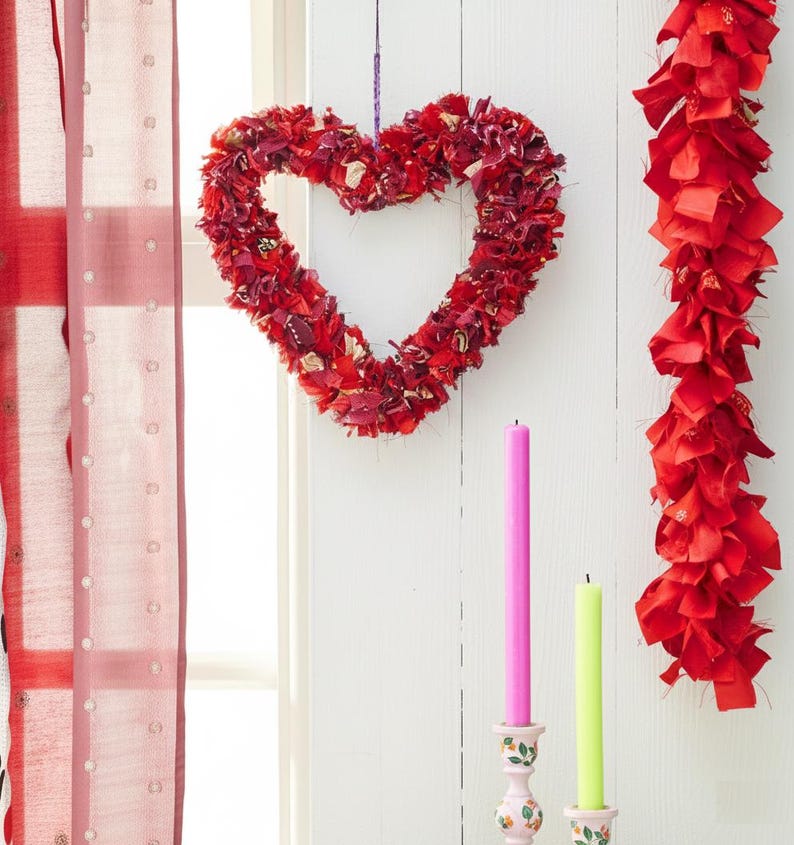 May include: A red heart-shaped wreath made of fabric hangs on a white wall. A red fabric garland is to the right. Two candles, one pink and one green, sit in decorative holders below.