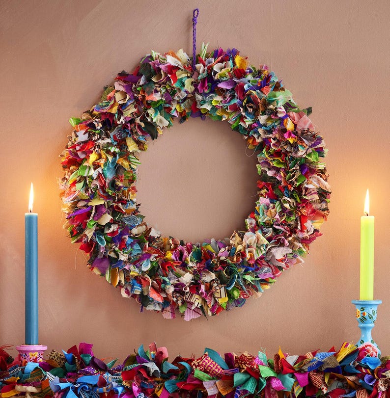 May include: A colorful, handmade wreath made of fabric scraps hangs on a wall. The wreath is a vibrant mix of red, green, blue, and yellow. Two lit candles flank the wreath, one blue and one green, adding a warm glow to the scene.