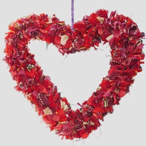 May include: A heart-shaped wreath made of red fabric scraps. The wreath is suspended by a purple string. The fabric pieces are various shades of red and burgundy, with some gold accents.