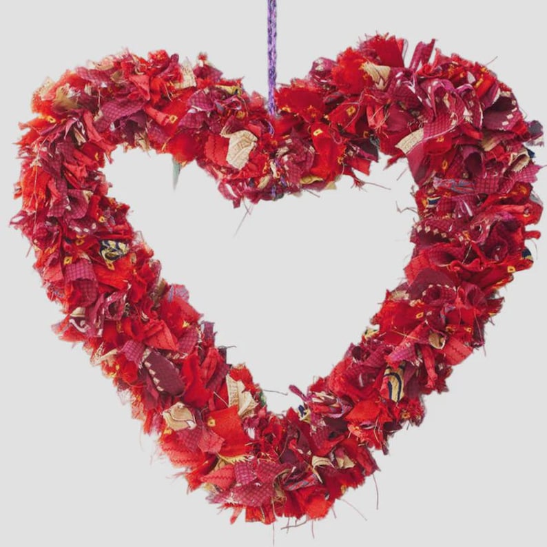 May include: A heart-shaped wreath made of red fabric scraps. The wreath is suspended by a purple string. The fabric pieces are various shades of red and burgundy, with some gold accents.