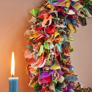 May include: A colorful, handmade wreath composed of fabric scraps in various colors, including red, green, yellow, and purple. A blue candle with a lit flame is in the foreground, set against a neutral background.
