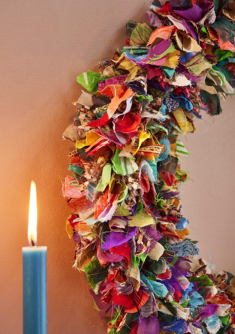 May include: A colorful, handmade wreath composed of fabric scraps in various colors, including red, green, yellow, and purple. A blue candle with a lit flame is in the foreground, set against a neutral background.