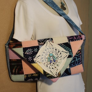 May include: A patchwork fabric bag with a crocheted embellishment and a denim strap. The bag is a light pink, blue, and yellow color with a floral pattern. The bag is being worn by a white shirt.
