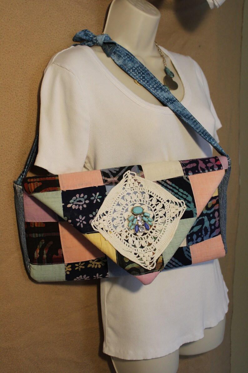 May include: A patchwork fabric bag with a crocheted embellishment and a denim strap. The bag is a light pink, blue, and yellow color with a floral pattern. The bag is being worn by a white shirt.