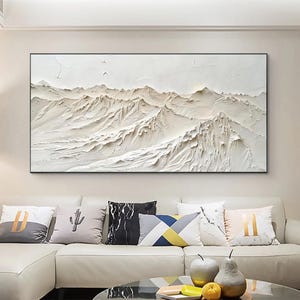 May include: A large, rectangular, textured white mountain landscape painting in a black frame. The artwork depicts a detailed relief of mountain ranges with varying shades of white and beige. The painting hangs above a white sofa with decorative pillows.