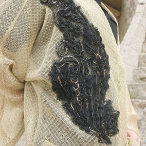May include: A beige cardigan with a black and gold embroidered design on the front. The cardigan has a pocket on the front and a loose fit.
