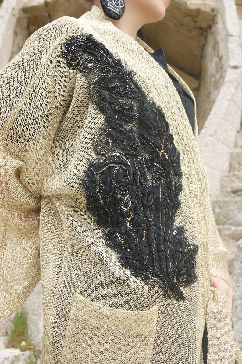May include: A beige cardigan with a black and gold embroidered design on the front. The cardigan has a pocket on the front and a loose fit.
