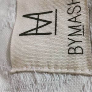 Pu&ograve; includere: Un'etichetta in tessuto bianco con testo nero che recita "BYMASH" e un logo "M" stilizzato.