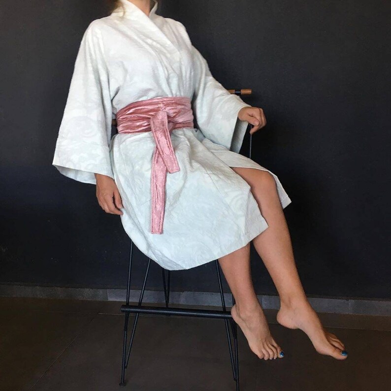 Pu&ograve; includere: Una veste bianca con una cintura di velluto rosa legata in vita. La veste &egrave; indossata da una persona seduta su uno sgabello di metallo nero.