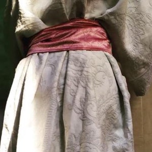 Pu&ograve; includere: Una veste bianca testurizzata con una cintura di raso rosa annodata in vita.