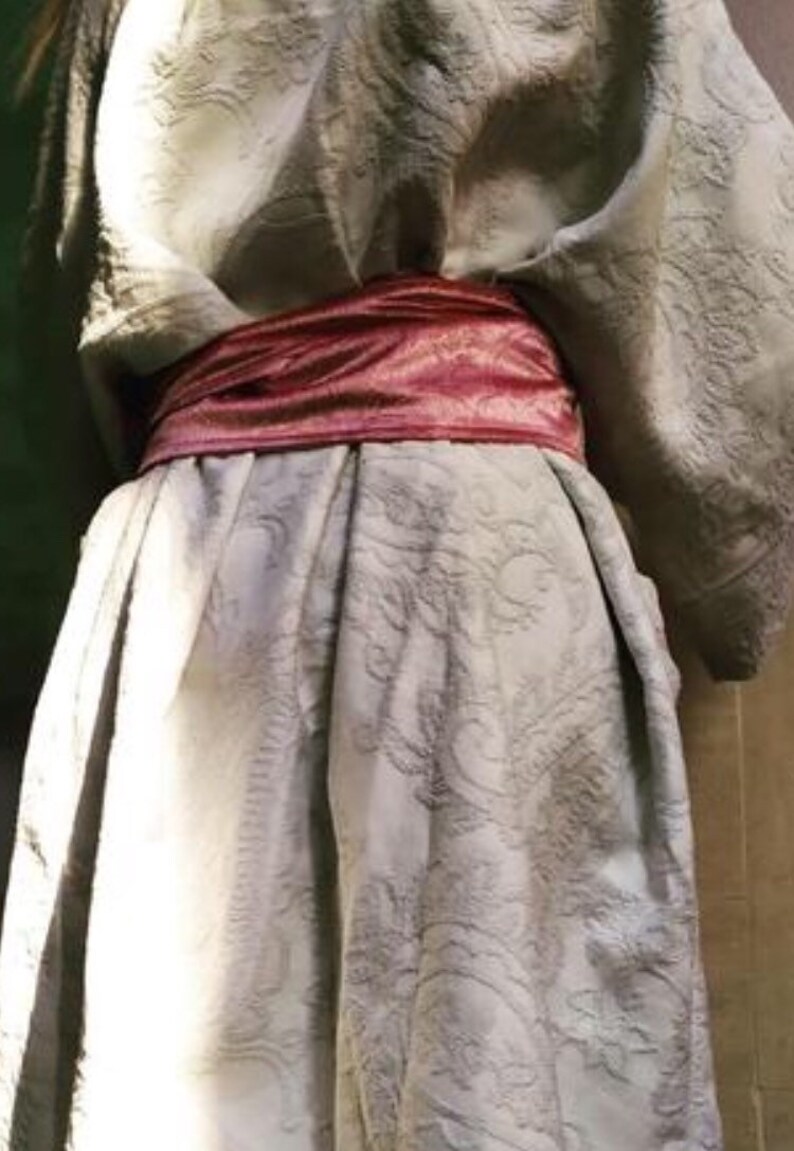 Pu&ograve; includere: Una veste bianca testurizzata con una cintura di raso rosa annodata in vita.