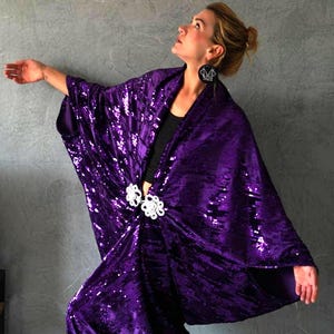 Può includere: Un kimono di paillettes viola con una chiusura decorativa bianca. Il kimono è realizzato in un tessuto morbido e fluido e ha una vestibilità rilassata.
