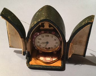 Puede incluir: Un pequeño reloj antiguo con una esfera blanca y agujas negras está alojado en una caja verde con bisagras en forma de libro.