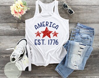 May include: White tank top with a red, white, and blue graphic that says "AMERICA EST. 1776" with three red stars. The tank top is laying on a wood surface with a pair of white sneakers, blue jeans, and sunglasses.