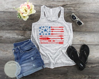 May include: A gray tank top with a red, white, and blue American flag design featuring stars and arrows. The tank top is on a wood background with denim shorts, sunglasses, and flip flops.