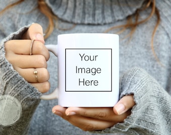 Könnte beinhalten: Eine weiße Keramiktasse mit einem schwarzen Rand um den Text "Your Image Here". Die Tasse wird von einer Person gehalten, die einen grauen Pullover trägt.