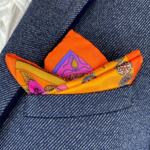 May include: A colorful pocket square with a geometric pattern in shades of orange, yellow, pink, purple, and blue. The pocket square is folded and placed in the pocket of a navy blue blazer.