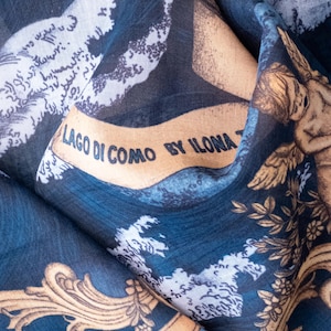 May include: A dark blue fabric featuring a printed design of golden cherubs, ornate scrollwork, and white cloud-like patterns. A tan banner across the center displays the black text LAGO DI COMO BY ILONA T, set against a deep blue background.