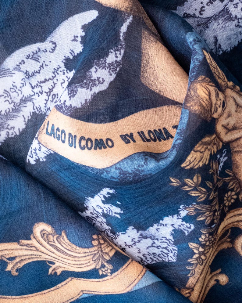 May include: A dark blue fabric featuring a printed design of golden cherubs, ornate scrollwork, and white cloud-like patterns. A tan banner across the center displays the black text LAGO DI COMO BY ILONA T, set against a deep blue background.