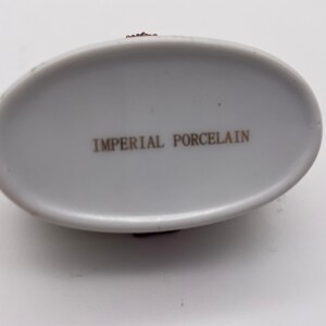 May include: The bottom of a white oval porcelain trinket box with the words "Imperial Porcelain" printed in gold.