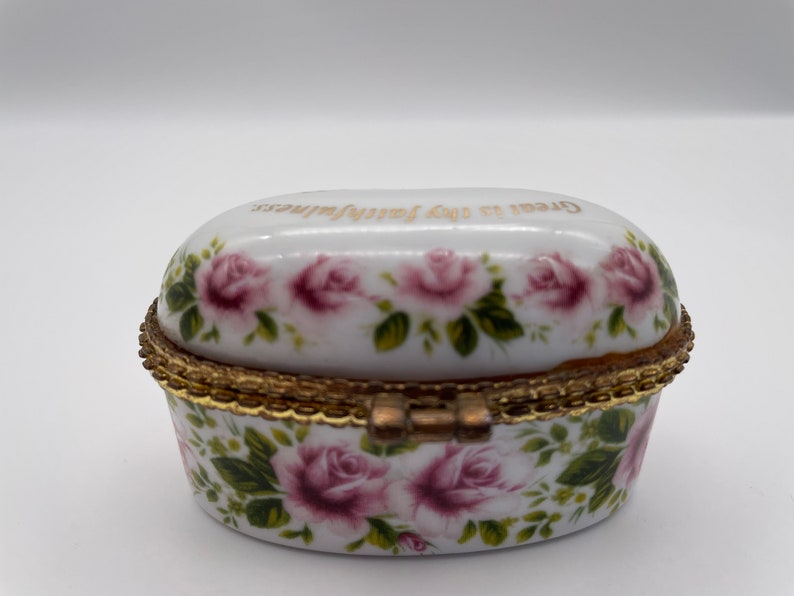May include: White porcelain trinket box with a gold trim and a hinged lid. The box is decorated with pink roses and green leaves. The lid has the words "Great is thy faithfulness" written in gold lettering.