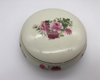 May include: A round, cream-colored ceramic trinket box with a lid. The lid is decorated with a cluster of pink and burgundy roses and green leaves. A thin gold band encircles the base and lid.