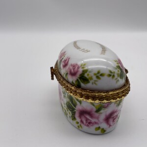 May include: Oval porcelain trinket box with a gold trim and floral design. The box is decorated with pink roses and green leaves. The lid has the inscription "Remember Me" in gold lettering.