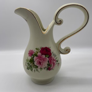 May include: A white ceramic pitcher with a gold trim and a floral design featuring pink and red roses. The pitcher has a curved handle.