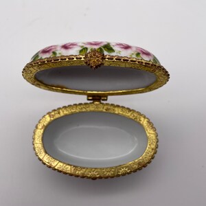 May include: Oval porcelain trinket box with a floral design and gold trim. The lid is open and the inside is white.